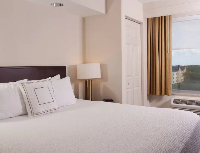 Bright and cozy king room at SpringHill Suites by Marriott featuring a bed with white linens, accent pillow, bedside lamp, and a window with beige curtains overlooking the outside.