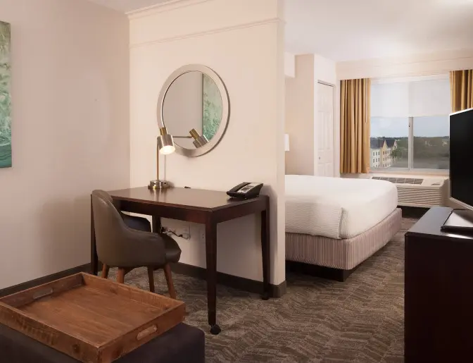 Modern king guest room at SpringHill Suites by Marriott featuring a bed, work desk with a lamp and phone, round mirror, decorative wall art, and a window with beige drapes.