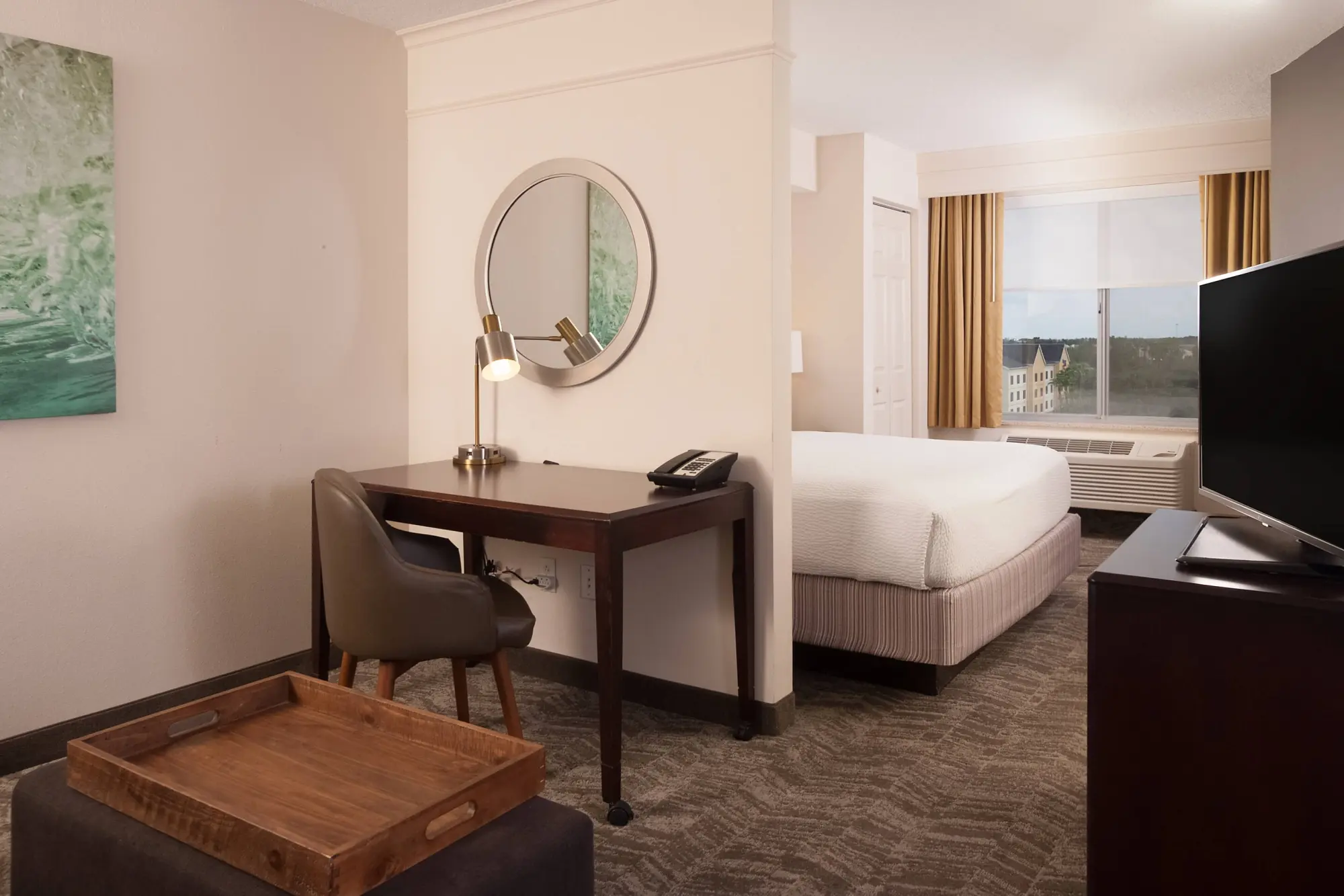 Modern king guest room at SpringHill Suites by Marriott featuring a bed, work desk with a lamp and phone, round mirror, decorative wall art, and a window with beige drapes.