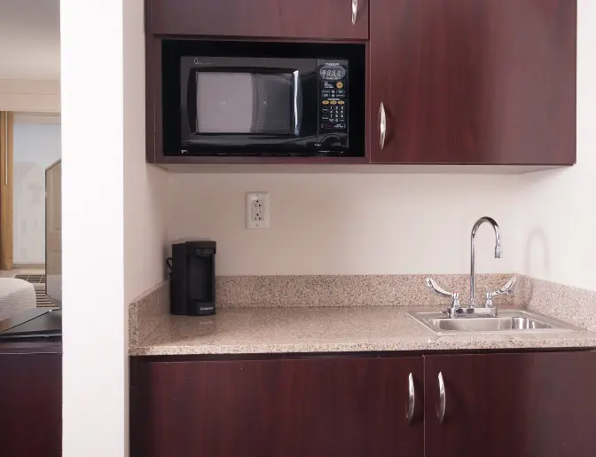 Kitchenette area in a guest room at SpringHill Suites by Marriott featuring a microwave, small sink, granite countertop, and a coffee maker, with dark wood cabinets.