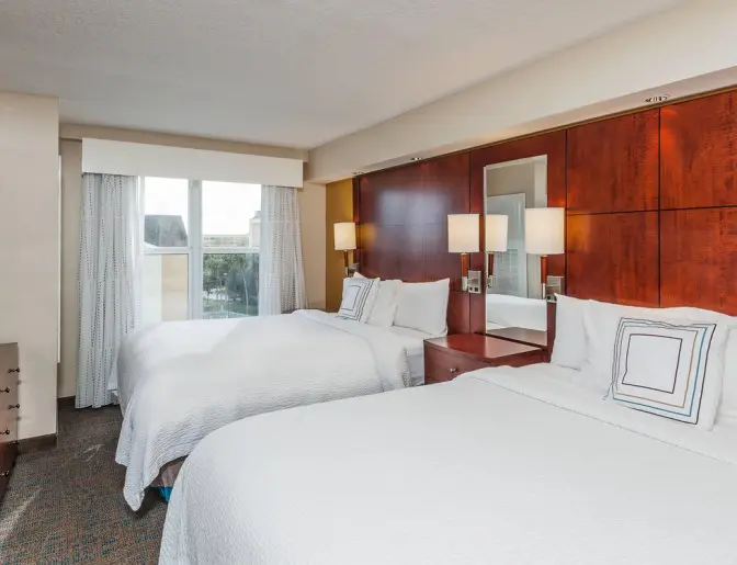 Hotel room at Residence Inn by Marriott with a two queen beds, white linens, a wooden headboard, bedside tables with lamps, and a window offering a view of the nearby area.