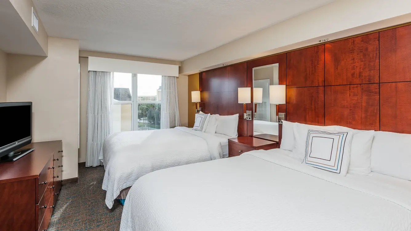 Hotel room at Residence Inn by Marriott with a two queen beds, white linens, a wooden headboard, bedside tables with lamps, and a window offering a view of the nearby area.