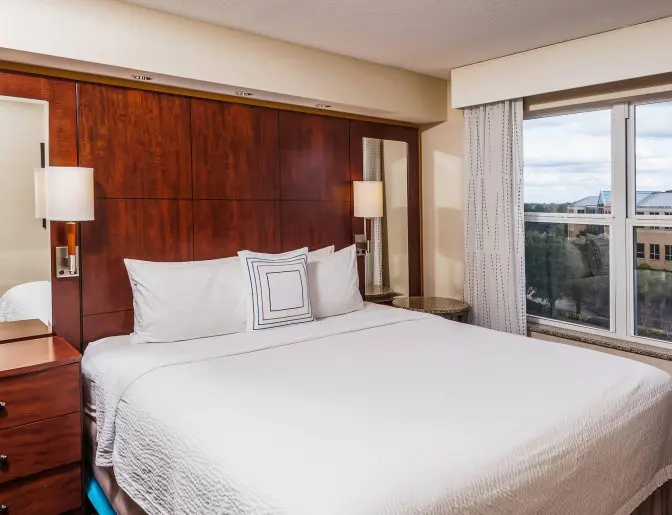 Hotel room at Residence Inn by Marriott with a king bed, white linens, a wooden headboard, bedside tables with lamps, and a window offering a view of the nearby area.
