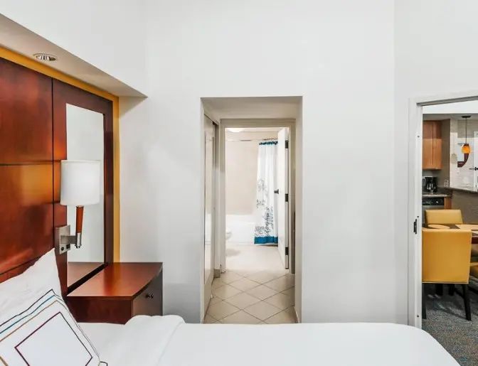 Hotel room featuring a king bed with white linens and a wooden headboard, a doorway leading to a bathroom with a shower curtain, and a dining area with yellow chairs and a kitchenette.
