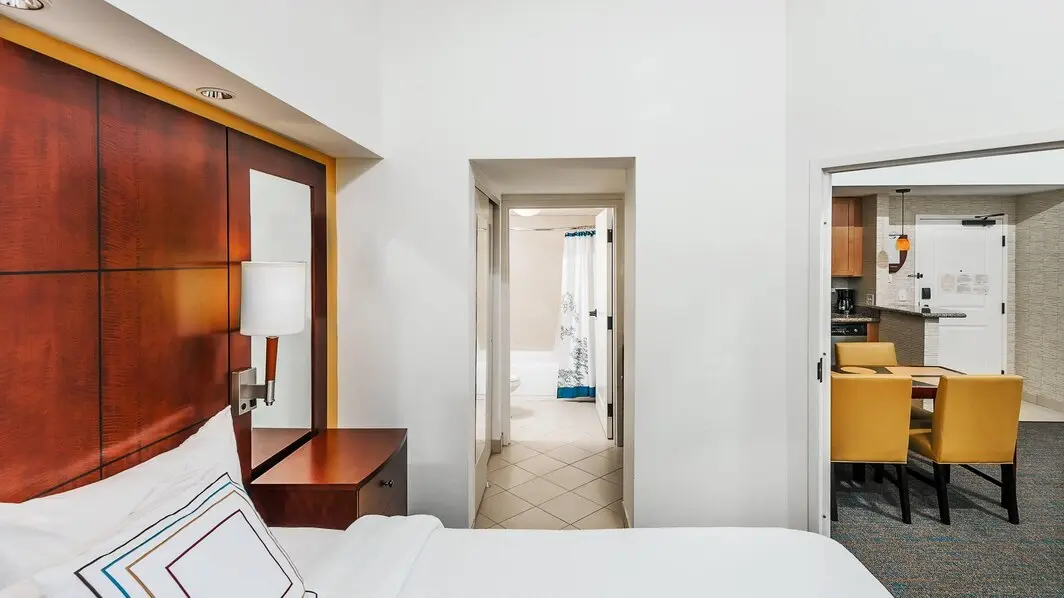 Hotel room featuring a king bed with white linens and a wooden headboard, a doorway leading to a bathroom with a shower curtain, and a dining area with yellow chairs and a kitchenette.