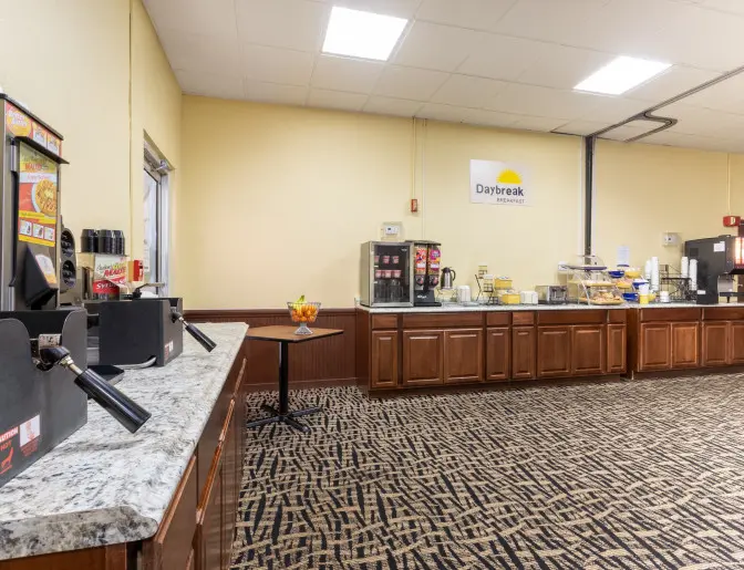 Breakfast area at the Days Inn Titusville hotel featuring a variety of food and beverage options including a waffle maker, coffee machine, cereal dispensers, and baked goods.