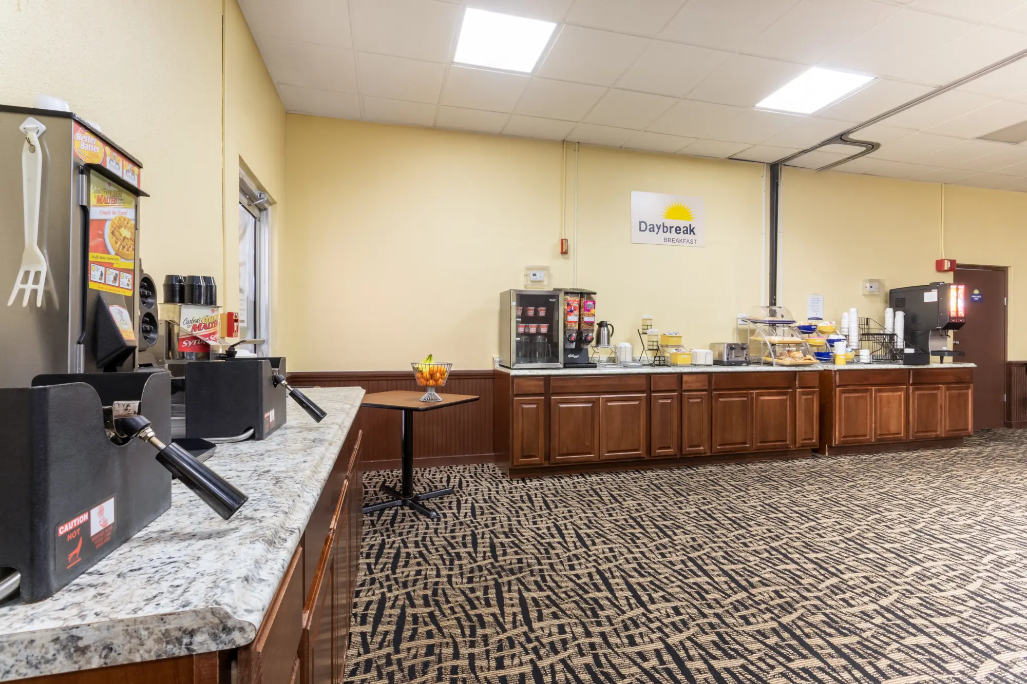 Breakfast area at the Days Inn Titusville hotel featuring a variety of food and beverage options including a waffle maker, coffee machine, cereal dispensers, and baked goods.
