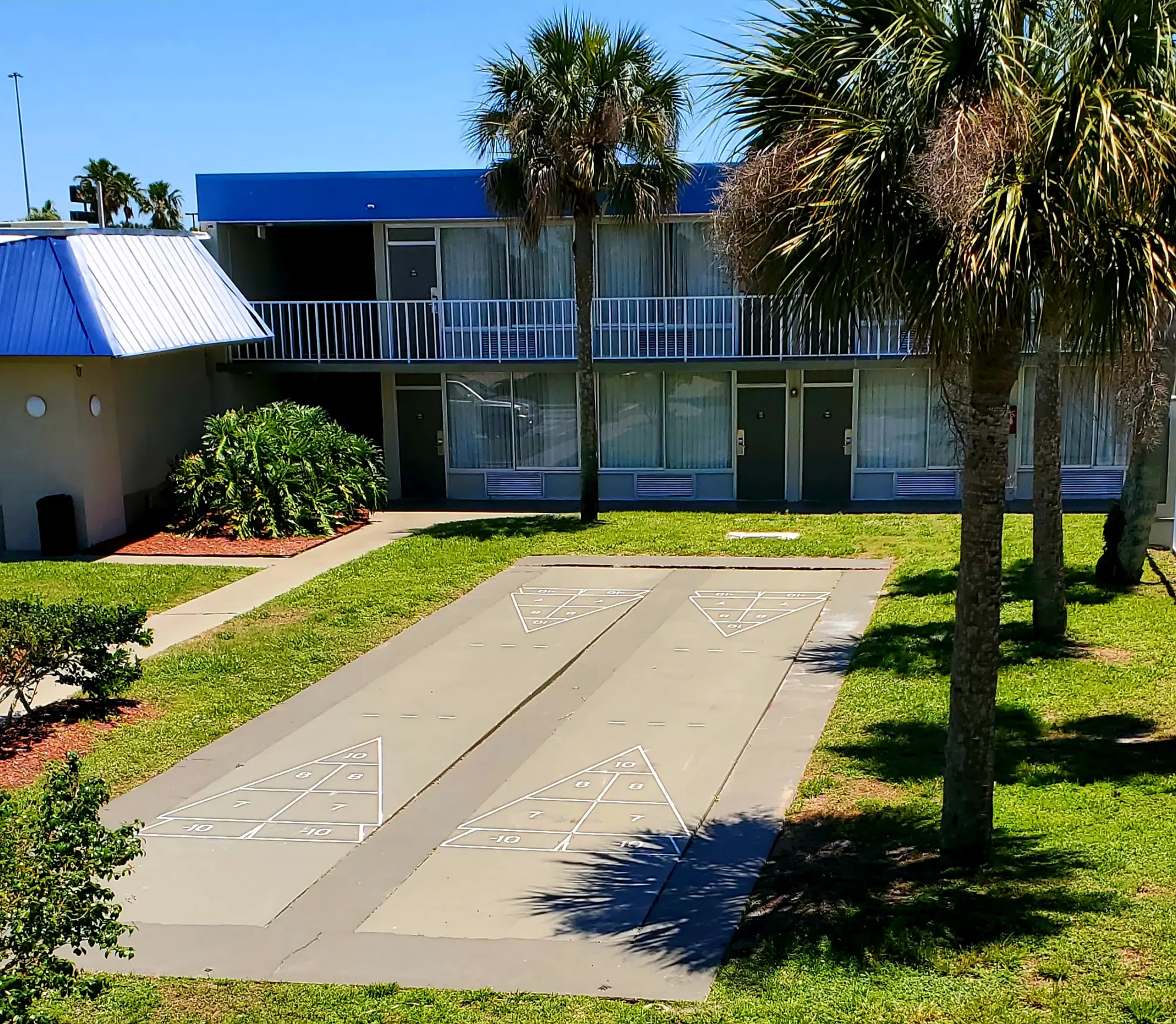 Days Inn Titusville hotel outdoor activity area featuring a shuffleboard court, palm trees, and well-maintained landscaping, with the hotel rooms visible in the background.