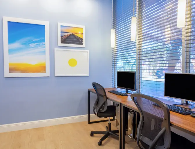Business center at the Days Inn Titusville hotel featuring two computer workstations with ergonomic chairs, located by a large window with blinds and decorated with framed artwork.