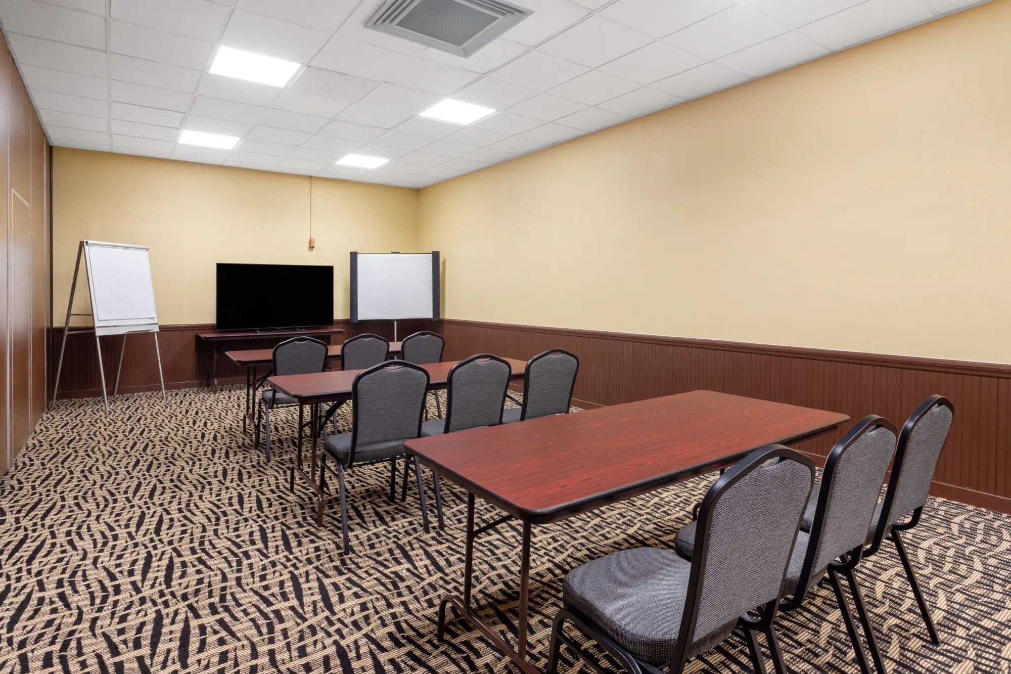 Meeting room at the Days Inn Titusville hotel featuring a large flat-screen TV, flip chart, whiteboard, and several tables with chairs arranged for a presentation.