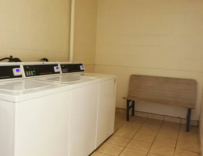 Laundry room at the Days Inn Titusville hotel featuring several commercial washers and a bench, with tiled floors and beige walls.