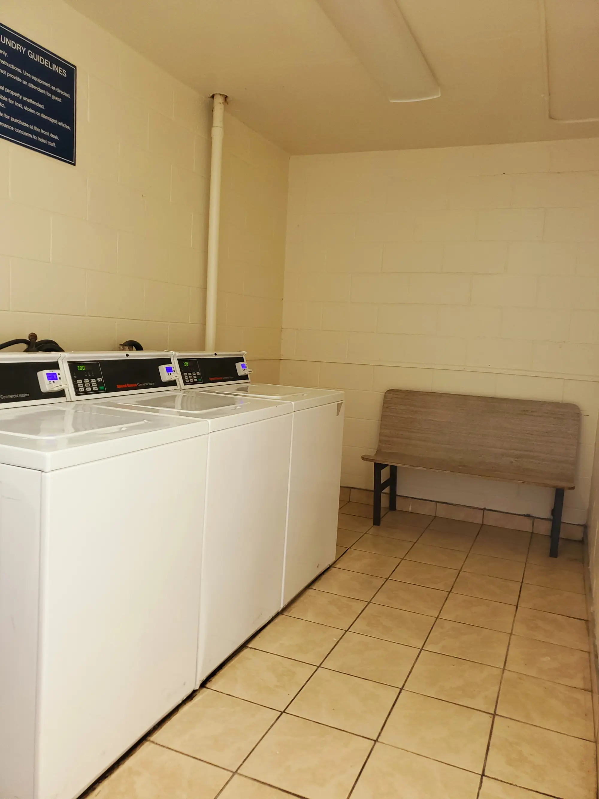 Laundry room at the Days Inn Titusville hotel featuring several commercial washers and a bench, with tiled floors and beige walls.