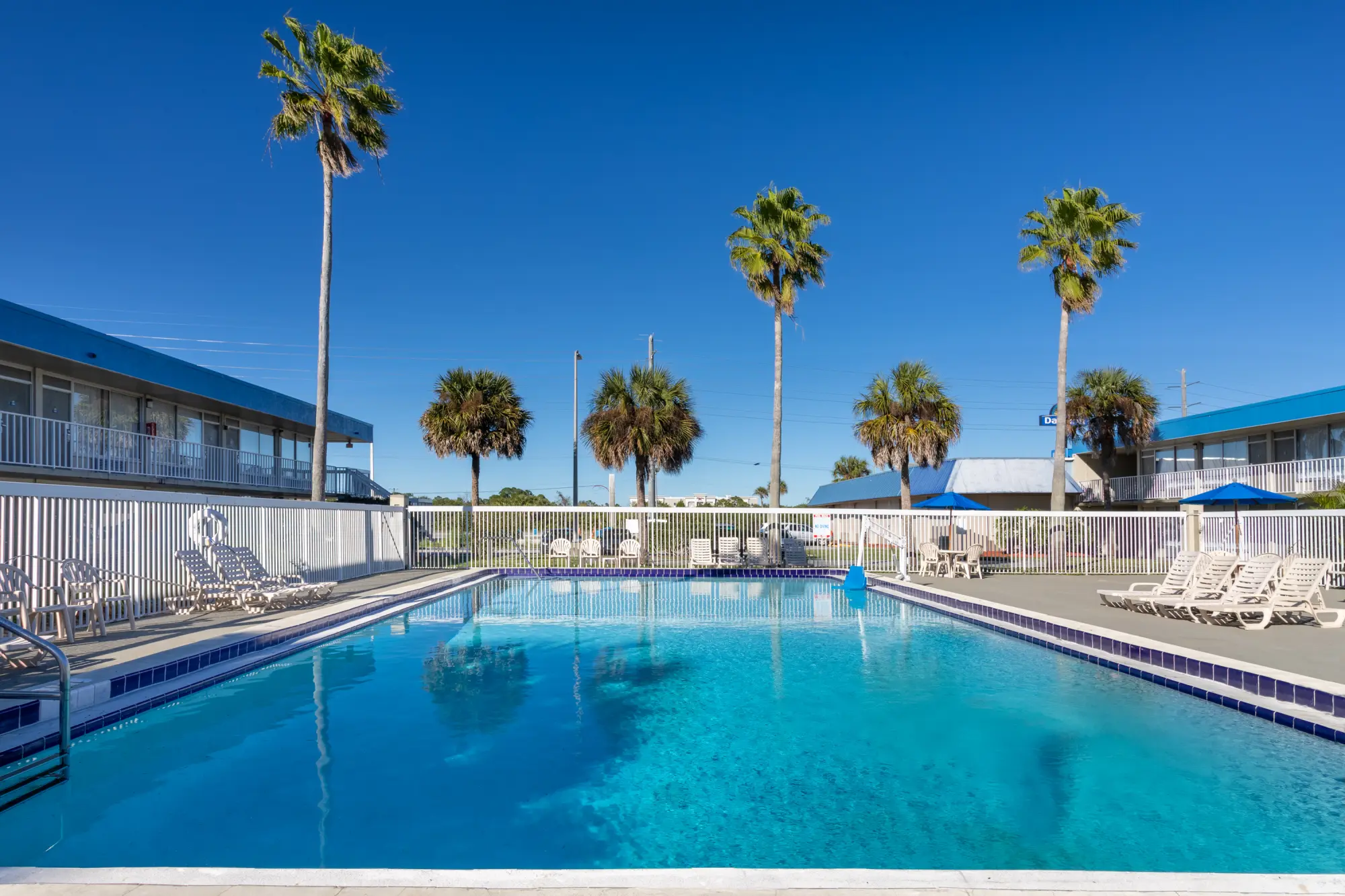 Outdoor swimming pool at the Days Inn Titusville hotel surrounded by lounge chairs and tall palm trees under a clear blue sky.