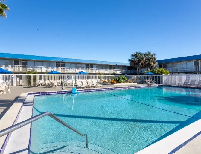 Large outdoor swimming pool at the Days Inn Titusville hotel with clear blue water, surrounded by lounge chairs, tables with blue umbrellas, and palm trees under a bright blue sky.