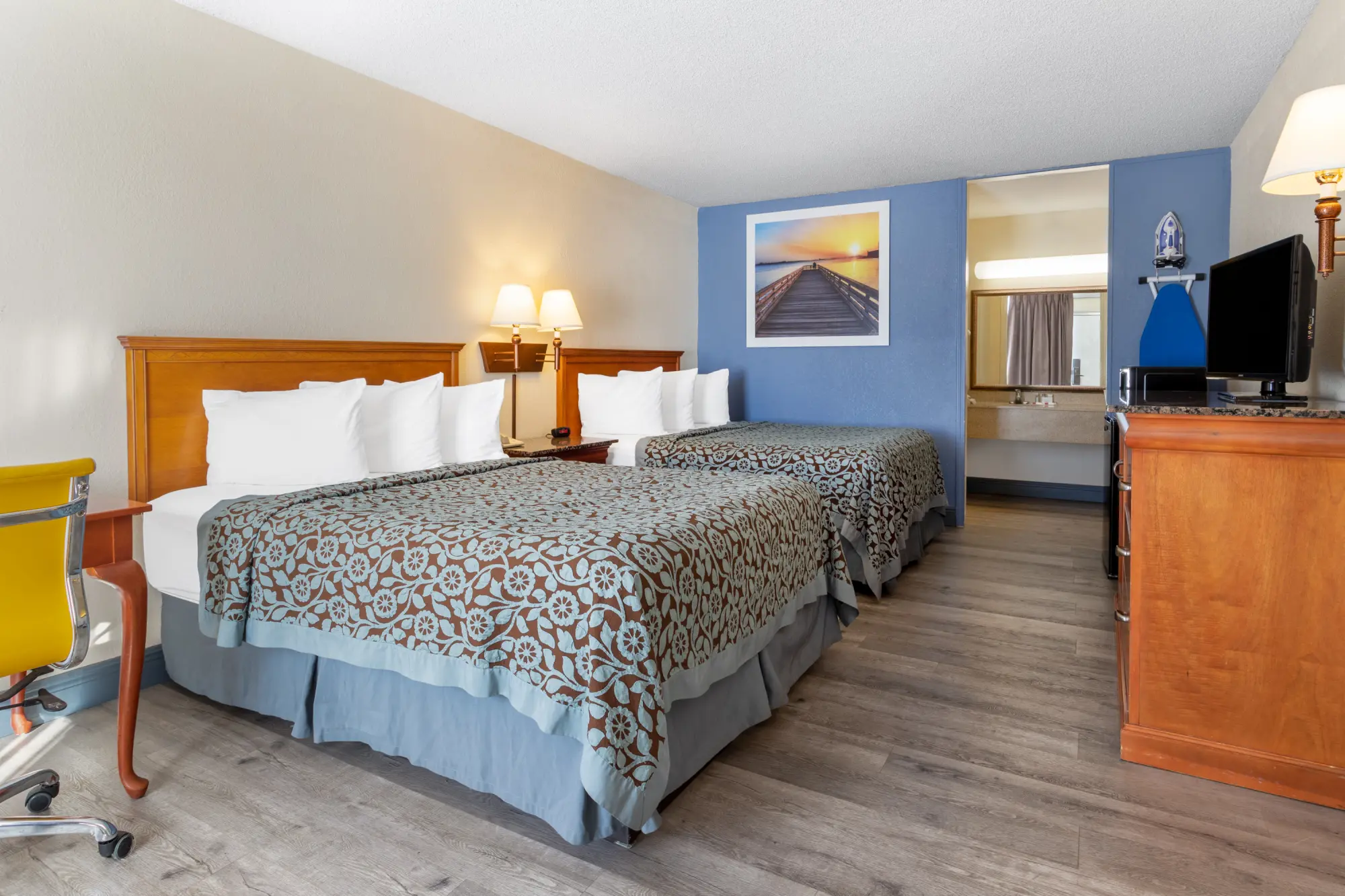 Guest room at Days Inn Titusville with two queen beds, a flat-screen TV, and a work desk. The room has a patterned bedspread, wooden furnishings, and a sunset boardwalk picture on a blue accent wall.