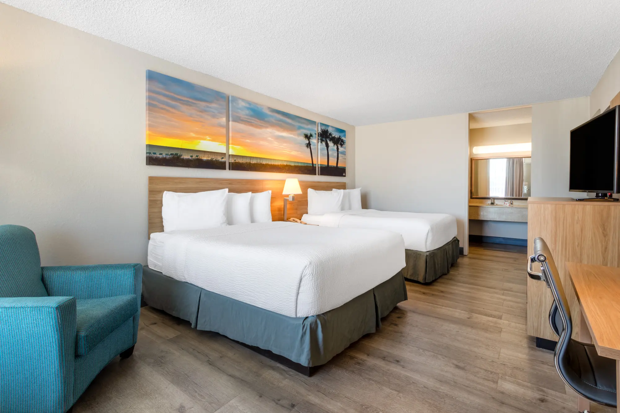 Guest room at Days Inn Titusville showcasing two queen-sized beds with white linens, a teal armchair, and wooden flooring. The room includes a flat-screen TV, a work desk, and a vanity area with a large mirror.