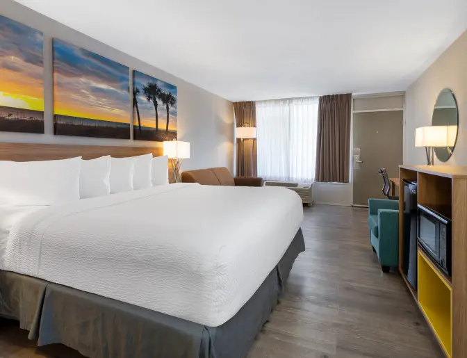 Spacious guest room at Days Inn Titusville with a king sized bed, white linens, and a large beach sunset artwork above the headboard. The room is furnished with a teal armchair, a work desk, and a wooden cabinet housing a flat-screen TV, mini-fridge, and microwave.