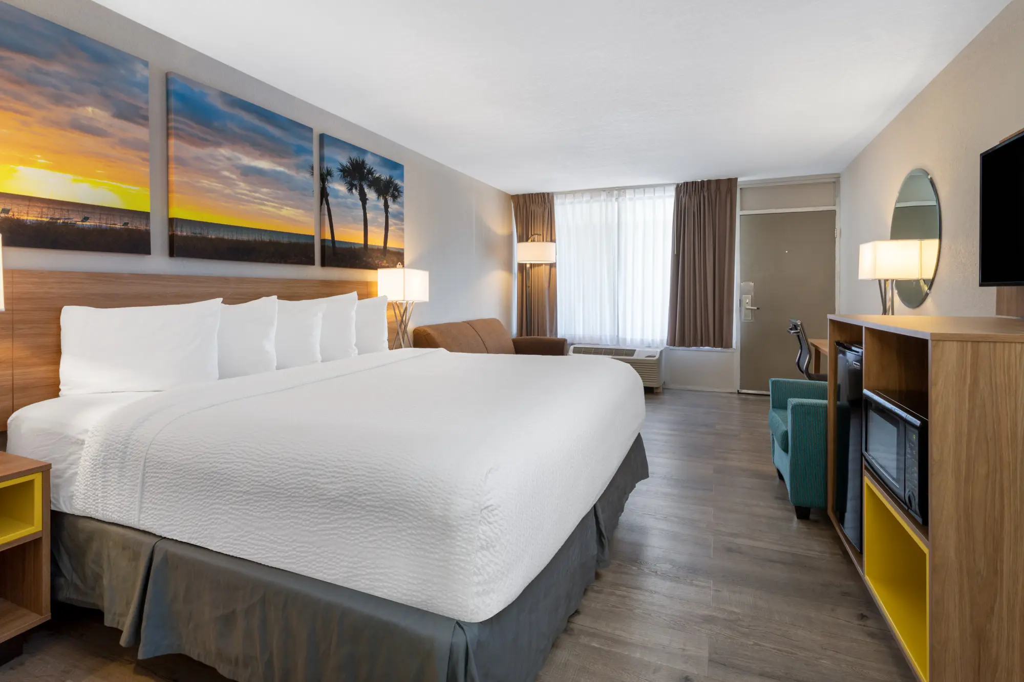 Spacious guest room at Days Inn Titusville with a king sized bed, white linens, and a large beach sunset artwork above the headboard. The room is furnished with a teal armchair, a work desk, and a wooden cabinet housing a flat-screen TV, mini-fridge, and microwave.