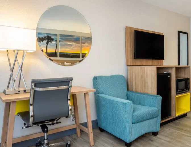 Comfortable guest room at Days Inn Titusville with a modern desk and lamp, a round mirror reflecting a sunset, a teal armchair, and a wooden cabinet with a flat-screen TV, mini-fridge, and microwave.