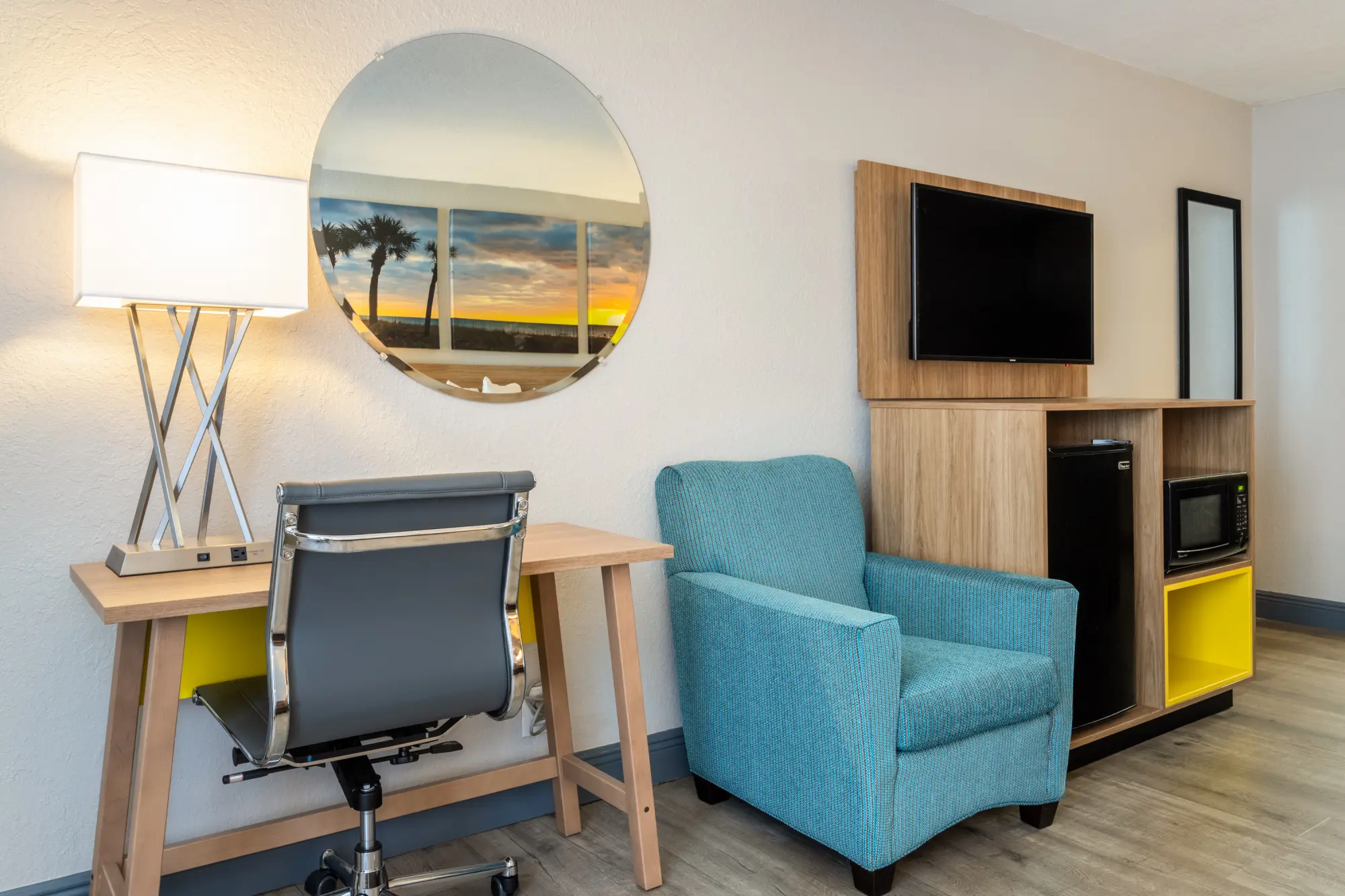 Comfortable guest room at Days Inn Titusville with a modern desk and lamp, a round mirror reflecting a sunset, a teal armchair, and a wooden cabinet with a flat-screen TV, mini-fridge, and microwave.