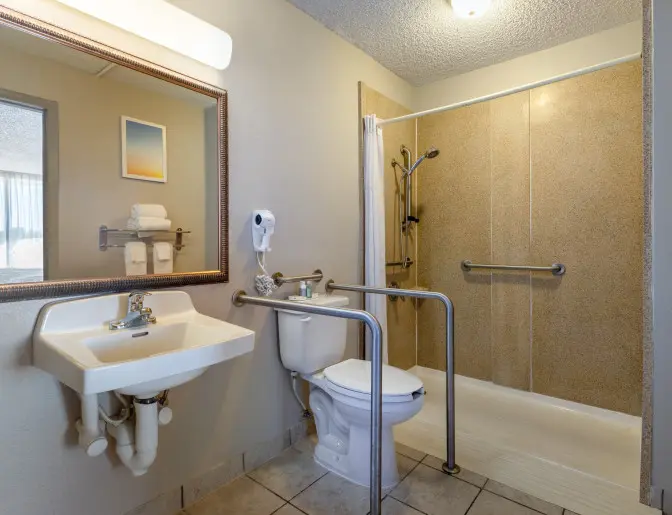 Accessible hotel bathroom featuring a walk-in shower with grab bars, a toilet with support rails, and a sink with exposed plumbing.