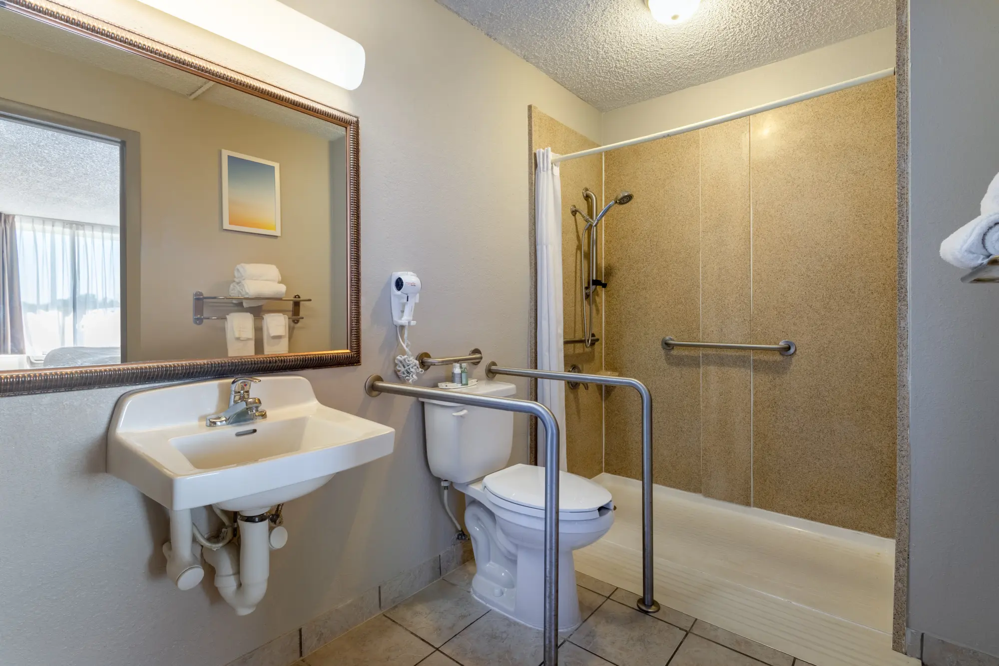 Accessible hotel bathroom featuring a walk-in shower with grab bars, a toilet with support rails, and a sink with exposed plumbing.