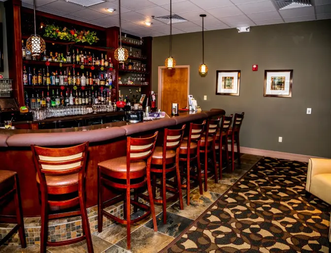 Hotel bar area with a row of wooden bar stools, a fully stocked bar, and hanging pendant lights.