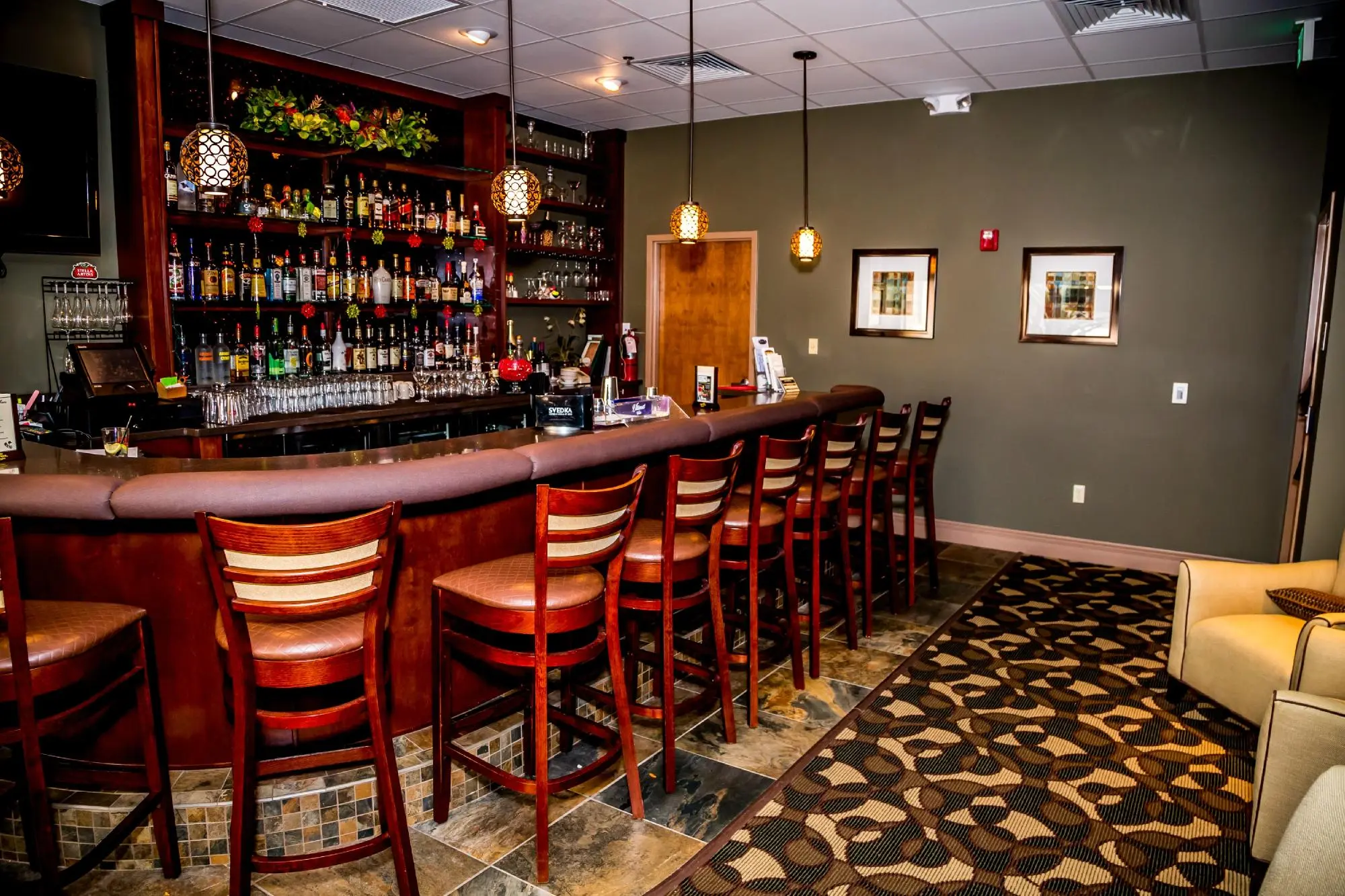 Hotel bar area with a row of wooden bar stools, a fully stocked bar, and hanging pendant lights.