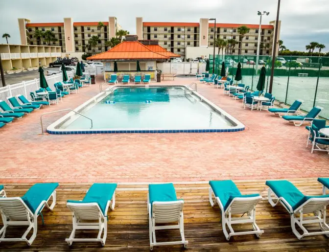 Ocean Landings Resort's outdoor pool surrounded by lounge chairs, tables with umbrellas, and a fenced tennis court in the background.