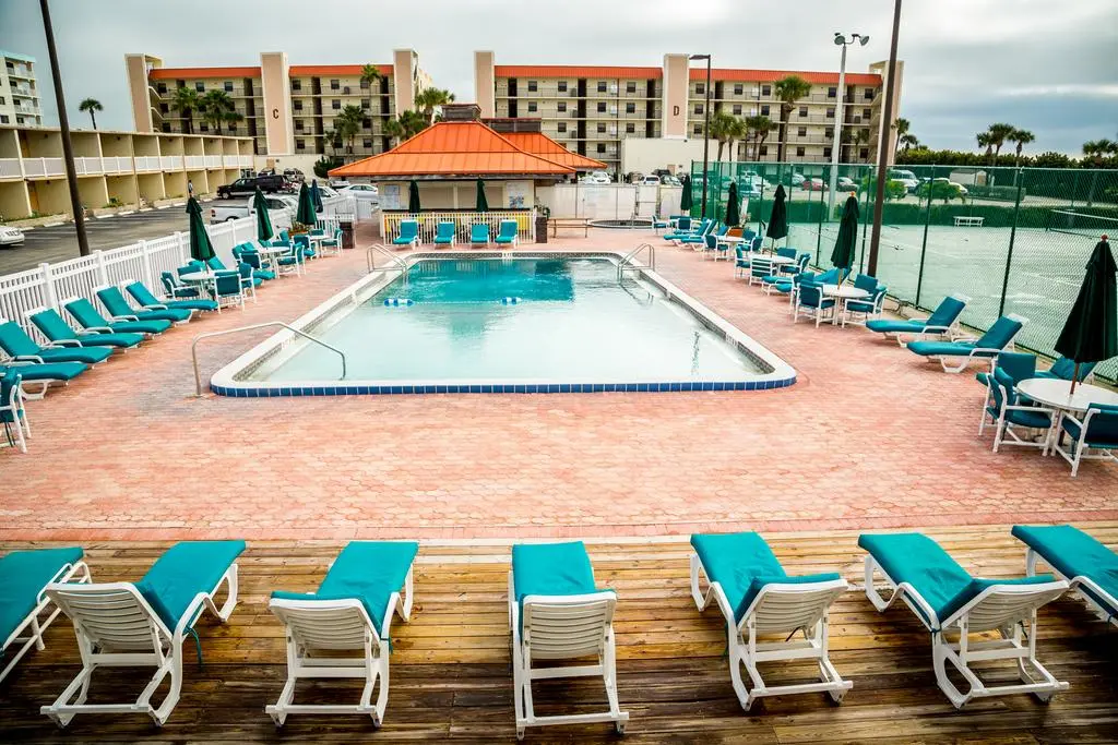 Ocean Landings Resort's outdoor pool surrounded by lounge chairs, tables with umbrellas, and a fenced tennis court in the background.