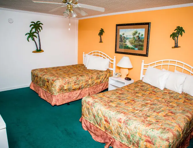 Hotel room with two double beds featuring colorful bedspreads, a ceiling fan, and tropical-themed decor on the walls.