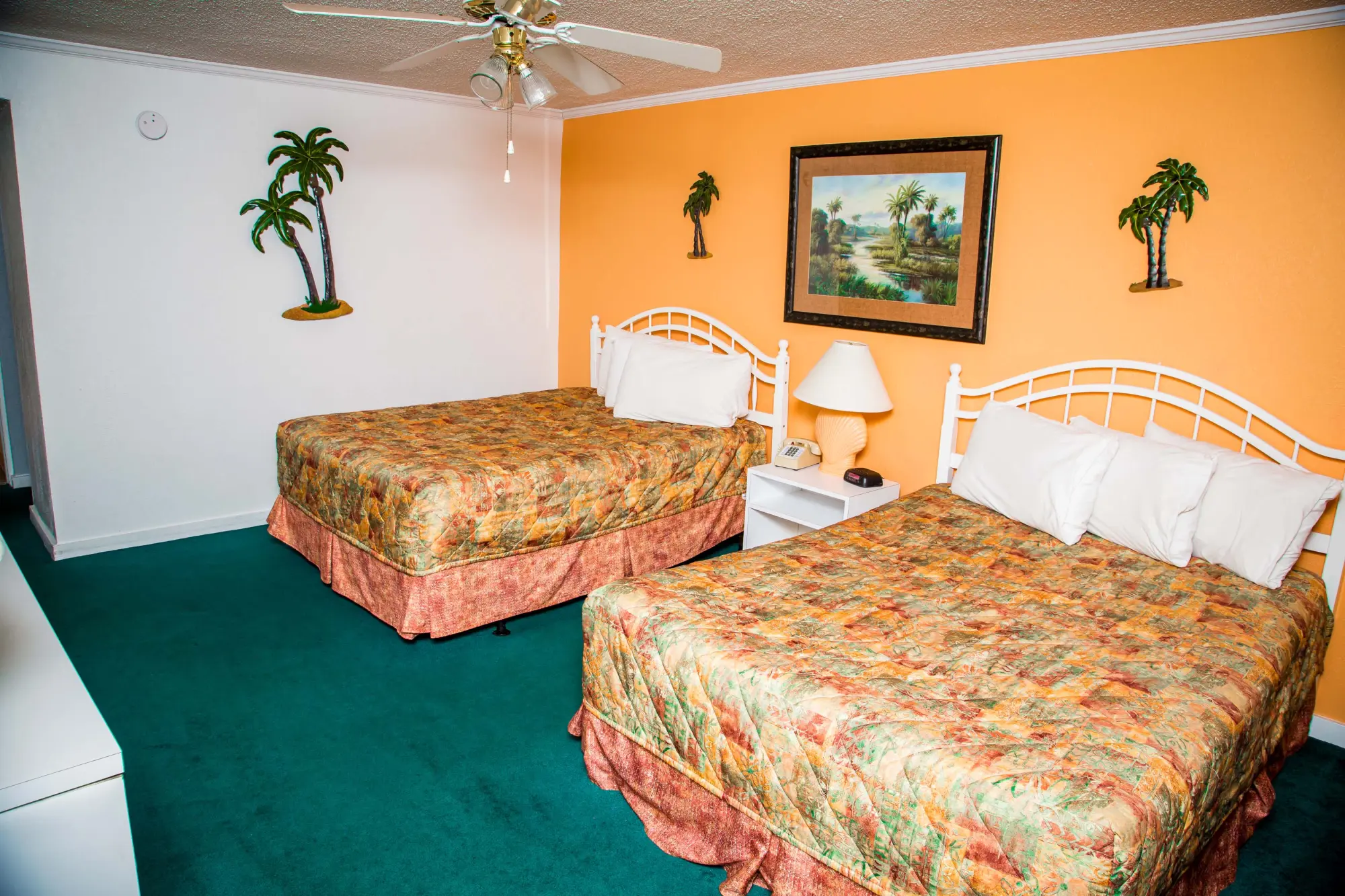 Hotel room with two double beds featuring colorful bedspreads, a ceiling fan, and tropical-themed decor on the walls.