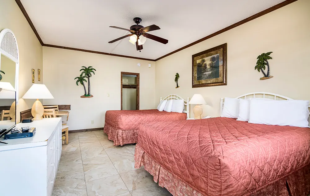 Hotel room with two queen beds, a ceiling fan, and tropical-themed decor including palm tree wall art and a framed painting.