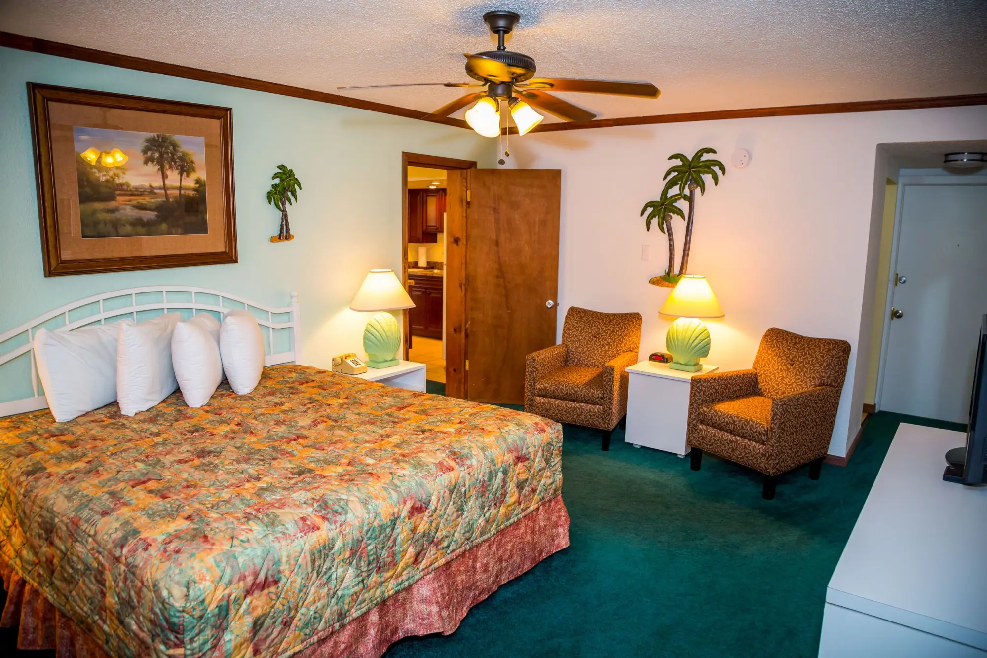 Hotel room featuring a king size bed with a colorful quilt, ceiling fan, and two armchairs with a small table and lamps.