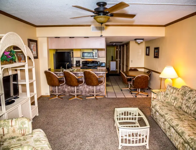 A spacious hotel suite living area featuring a kitchen with a breakfast bar and brown stools, a tropical-themed sofa, a wicker TV stand, and a dining table with chairs.