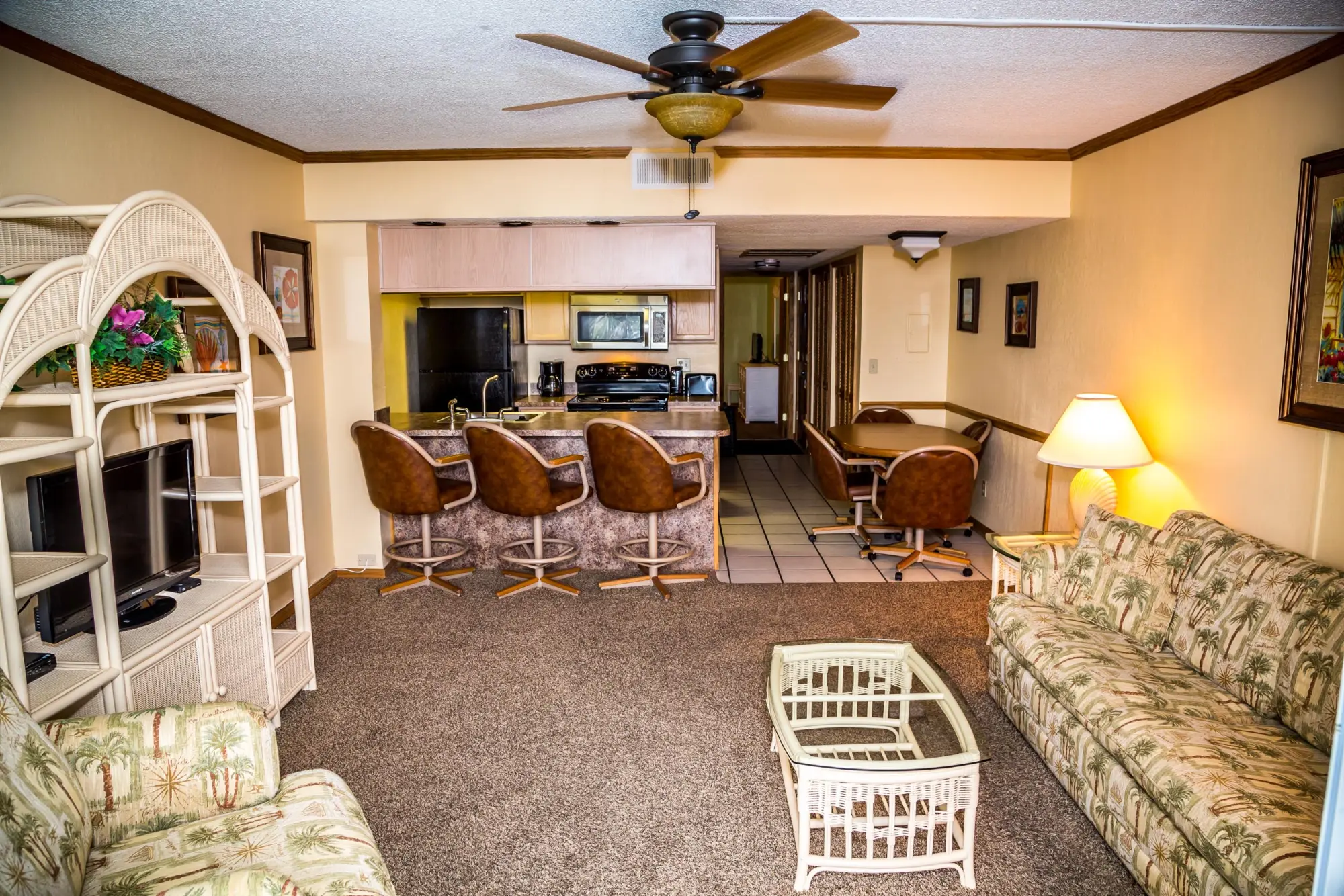 A spacious hotel suite living area featuring a kitchen with a breakfast bar and brown stools, a tropical-themed sofa, a wicker TV stand, and a dining table with chairs.