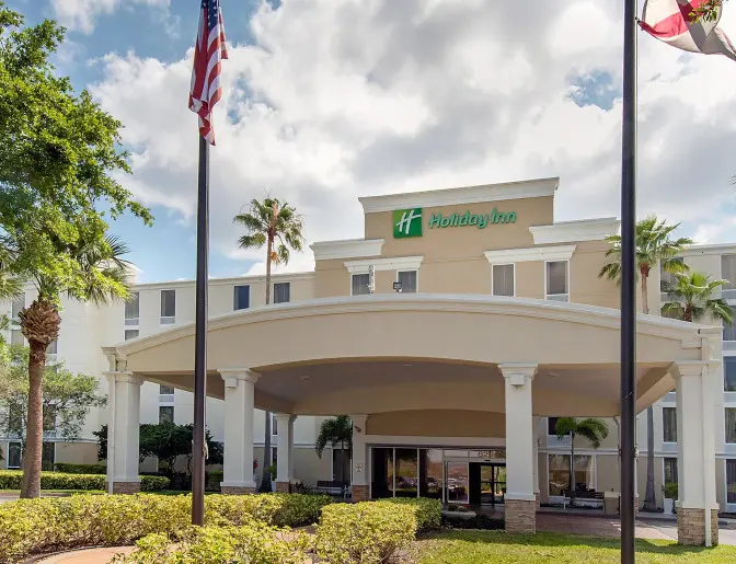Front entrance of a Holiday Inn hotel with a large arched canopy, American and state flags, and palm trees in the foreground.