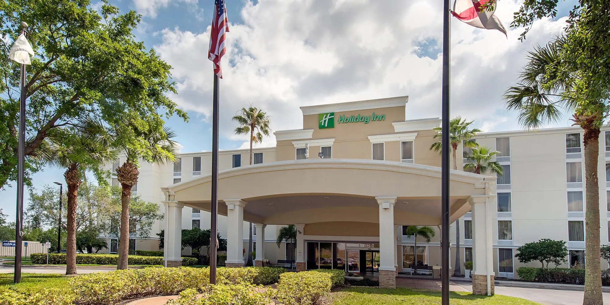 Front entrance of a Holiday Inn hotel with a large arched canopy, American and state flags, and palm trees in the foreground.