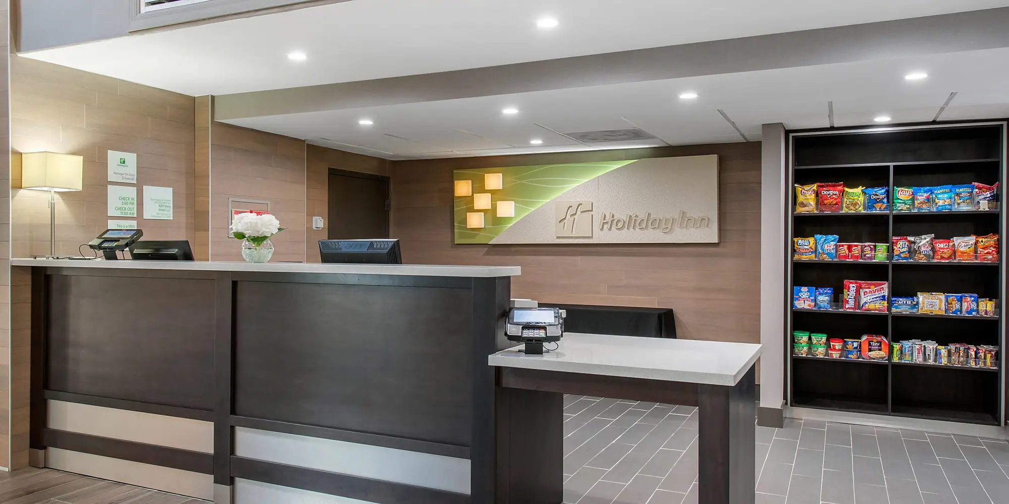 Holiday Inn hotel lobby with a modern front desk, flower arrangement, and a snack display shelf filled with assorted treats.