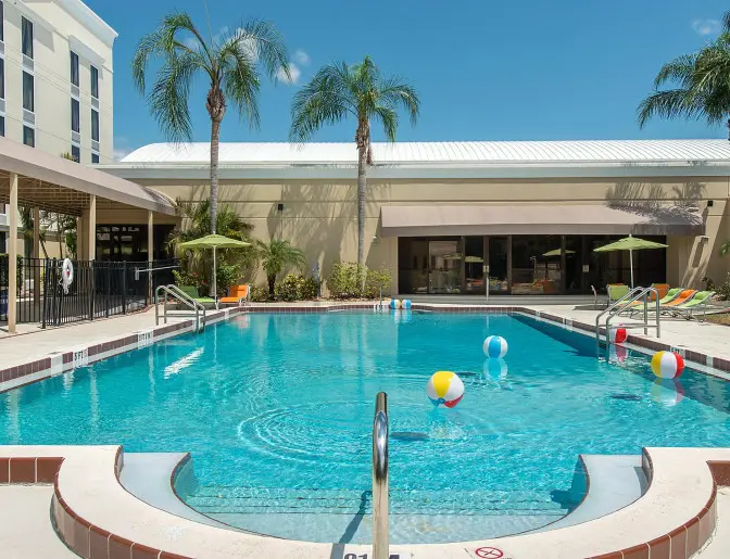 Outdoor swimming pool at Holiday Inn surrounded by palm trees, colorful lounge chairs, and beach balls floating in the water.