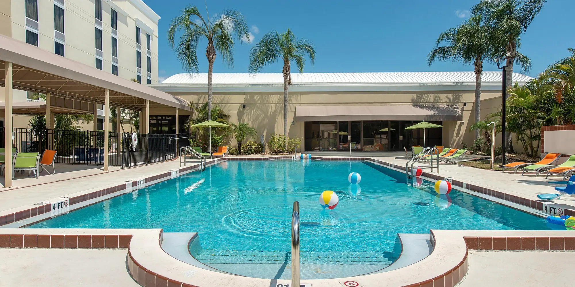 Outdoor swimming pool at Holiday Inn surrounded by palm trees, colorful lounge chairs, and beach balls floating in the water.