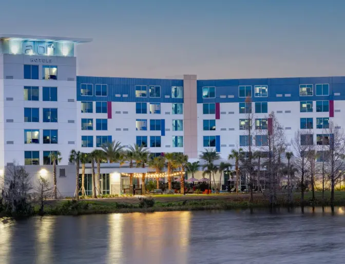 Aloft Orlando Lake Nona, a modern, multi-story hotel with a sleek, geometric design, featuring large windows and vibrant exterior lighting at dusk.