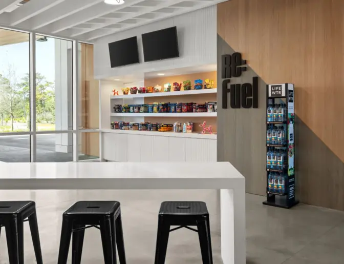 Snack area at Aloft Orlando Lake Nona with shelves full of snacks and drinks, a white countertop with black stools, and a LifeWTR display. Large windows allow natural light to brighten the area.