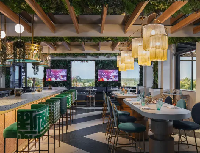 Stylish bar with green velvet stools, hanging plants, and modern gold accents, with large windows offering views of the surrounding landscape.