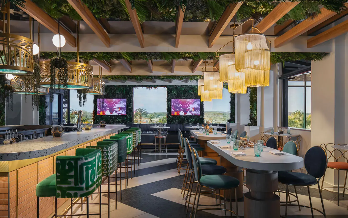 Stylish bar with green velvet stools, hanging plants, and modern gold accents, with large windows offering views of the surrounding landscape.