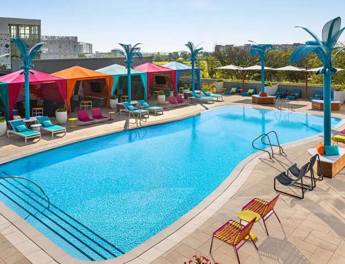 Vibrant pool area with colorful cabanas, bright lounge chairs, and unique blue palm tree sculptures surrounding a large outdoor pool.
