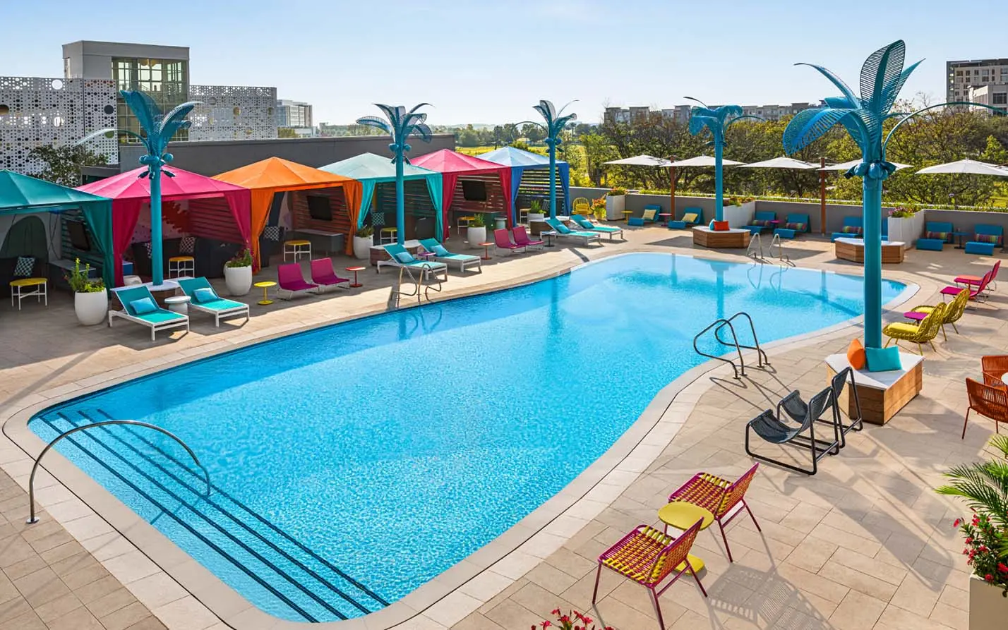 Vibrant pool area with colorful cabanas, bright lounge chairs, and unique blue palm tree sculptures surrounding a large outdoor pool.