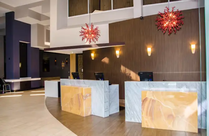 Modern reception area of Hilton Cocoa Beach featuring marble check-in desks with amber stone accents, warm wood-paneled walls, and contemporary coral-inspired light fixtures.