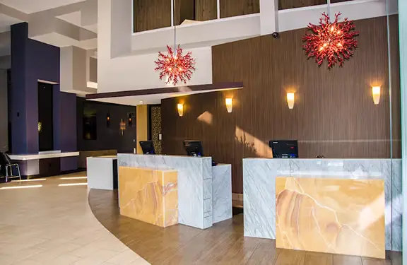 Modern reception area of Hilton Cocoa Beach featuring marble check-in desks with amber stone accents, warm wood-paneled walls, and contemporary coral-inspired light fixtures.