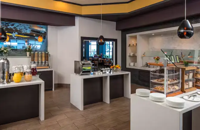 Hotel breakfast buffet at Hilton Cocoa Beach featuring a juice station, cereal dispensers, fresh pastries, a toaster, and neatly arranged dishware in a modern dining area.